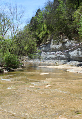 A massive blue stone wall along the edge of the Otter Creek frames the bend of the creek with vegetation.