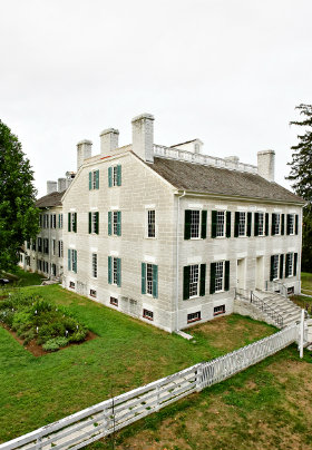 Historic white building with black shuttered windows, multiple chimneys and green gardens around.